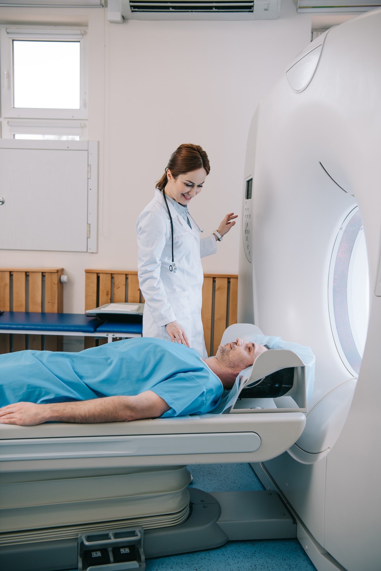 A healthcare professional assists a patient lying on a scanning bed, preparing for a medical imaging procedure in a clinical setting. The environment is bright and organized, indicating a focus on patient care.